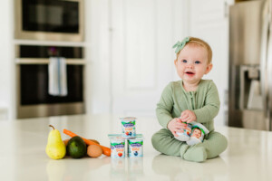 A baby wearing a green outfit and headband sits on a kitchen countertop, surrounded by fruit, vegetables, and containers of yogurt—perfect for starting solids with baby. In the background are a double oven with a cloth hanging on the handle and a stainless steel appliance.