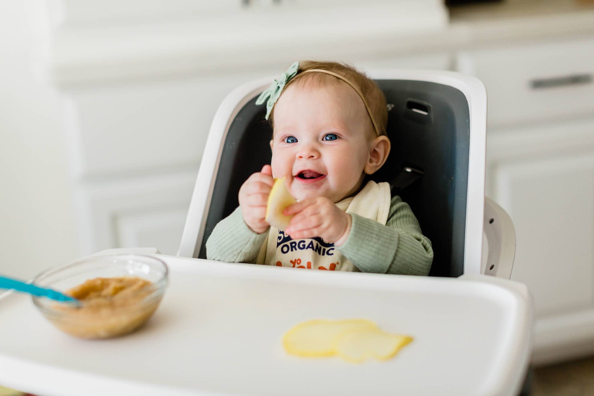 Baby girl sitting in a high chair holding a steamed slice of pear.
