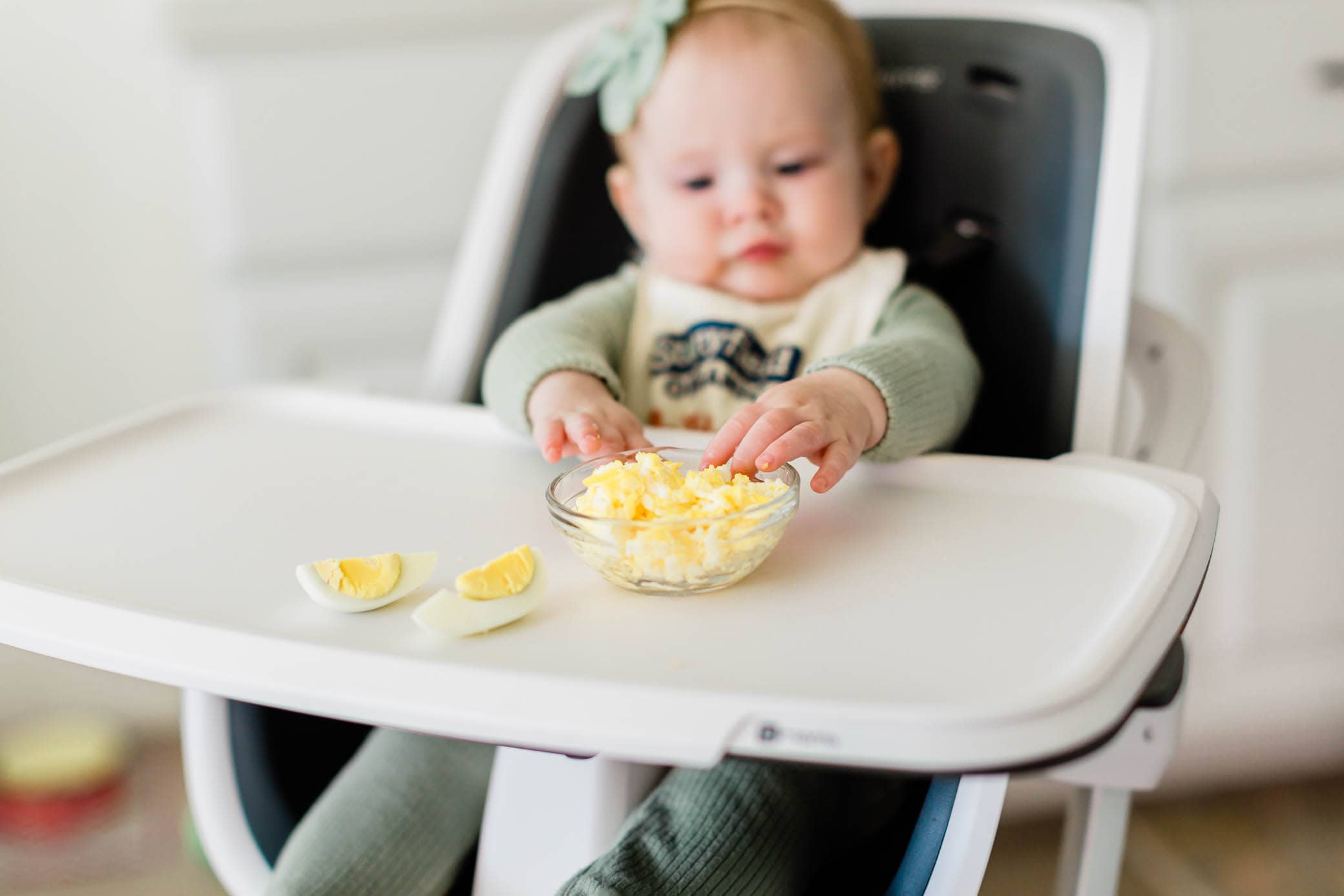 Baby girl sitting in a highchair reaching for the scrambled eggs.