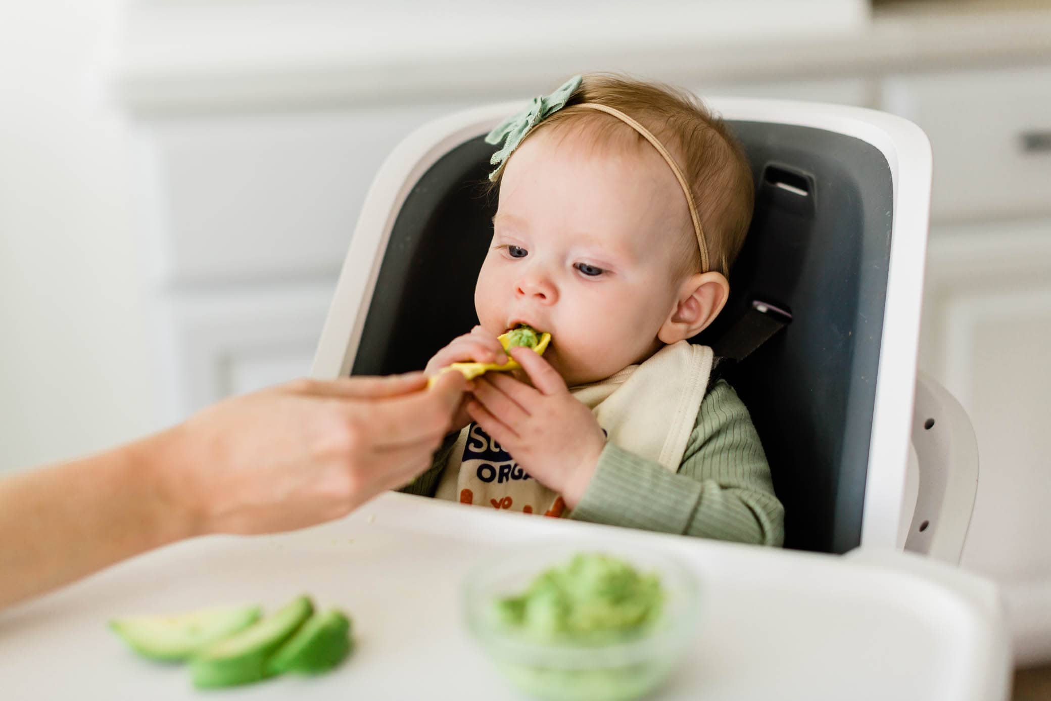 Baby girl sitting in a highchair being fed avocado.