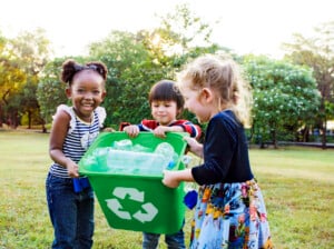 Three children stand outdoors holding a green recycling bin filled with plastic bottles.