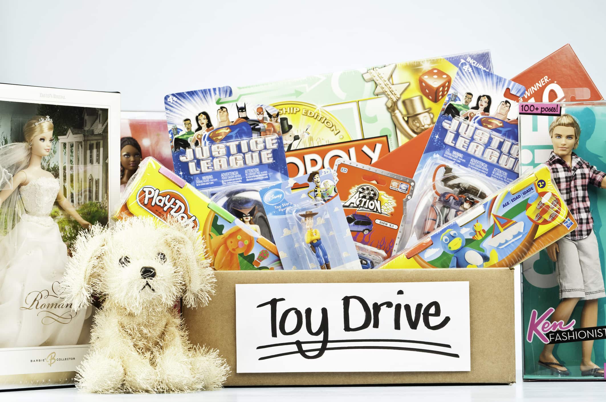 A large collection of toys and games in a cardboard box labelled for a holiday toy drive. Included in the box are Play-Doh, Justice League figurines, a Monopoly game, Scrabble game, a Disney Toy Story figurine, and an Action toy car. A cuddly toy dog sits in front of the box, and Barbie dolls and a Ken Fashionista doll flank the sides of the box.