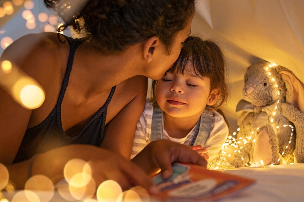 African american mother kissing her daughter on forehead while lying on bed in illuminated tent. Close up of mom kissing little cute girl with closed eyes holding story book in tent.