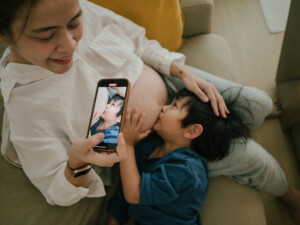 Pregnant mother and son sitting on the couch and son is kissing her pregnant belly and mother is taking a video by smart phone of it at home.