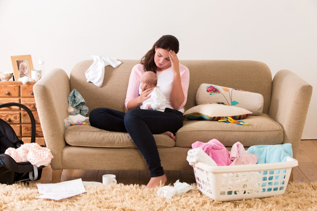 Mother with her baby on her knee. Her head is lowered as she sits on a sofa looking tired and stressed. The room looks untidy with clothes, nappies and other items scattered around the room.
