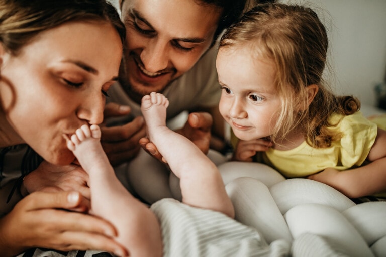 A woman kisses a baby's feet while a man and a young child, with blonde hair and wearing a yellow shirt, look on. The group is positioned closely together on a white surface, focused on the baby and welcoming the new sibling.
