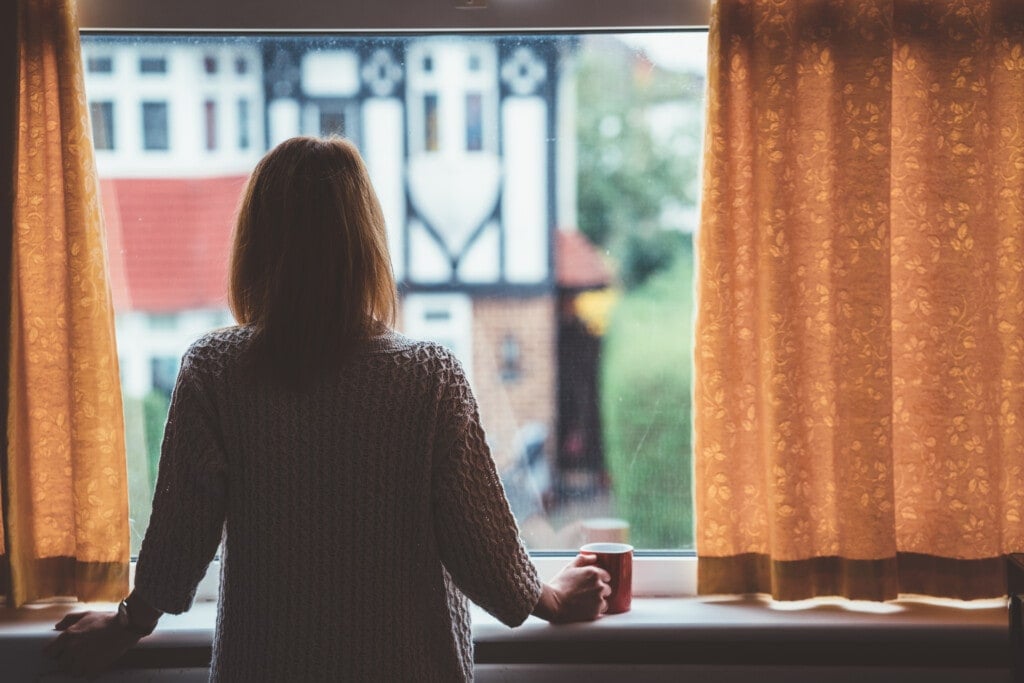 Rear view of woman at home staring through the window holding a cup of tea.