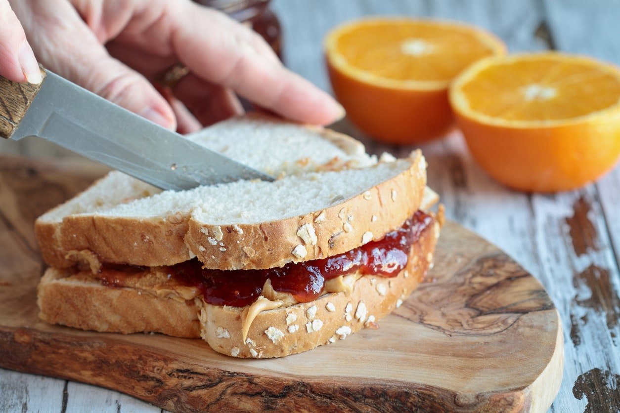 Womans hand cutting a peanut butter and Jelly sandwich on a rustic wooden cutting board.