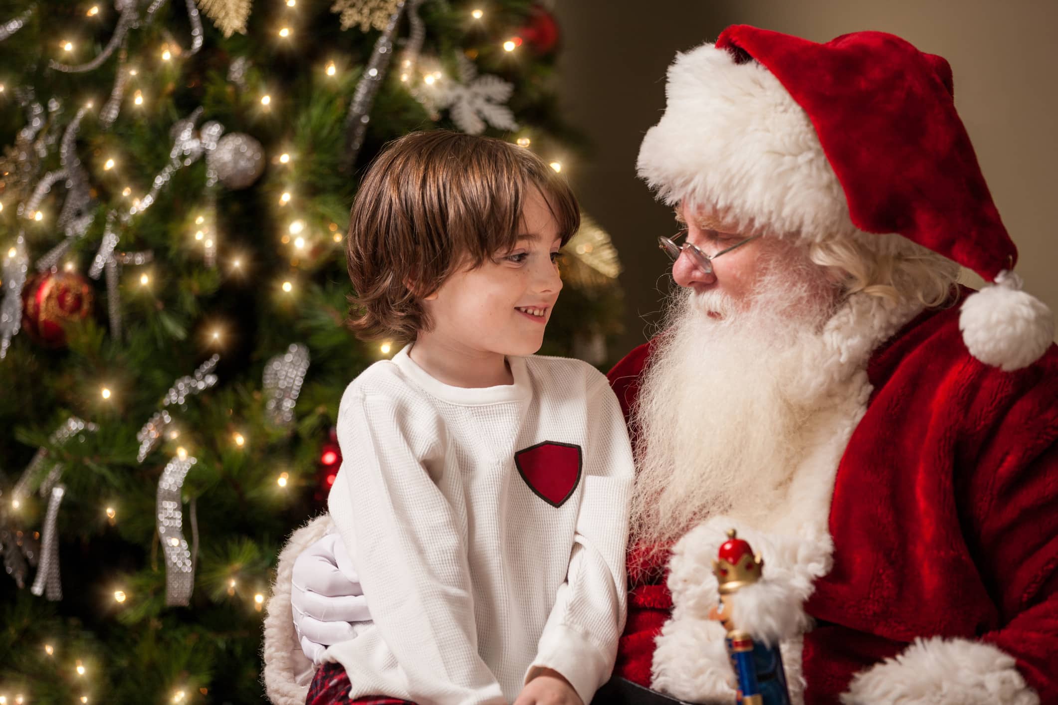 A young boy sits on Santa Claus' lap by a decorated Christmas tree in his living room. Santa has a real beard and the child looks at him with wonder, awe, and a hint of excitement.