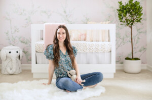 Naomi Coe sitting in front of a crib in a little girl's nursery.