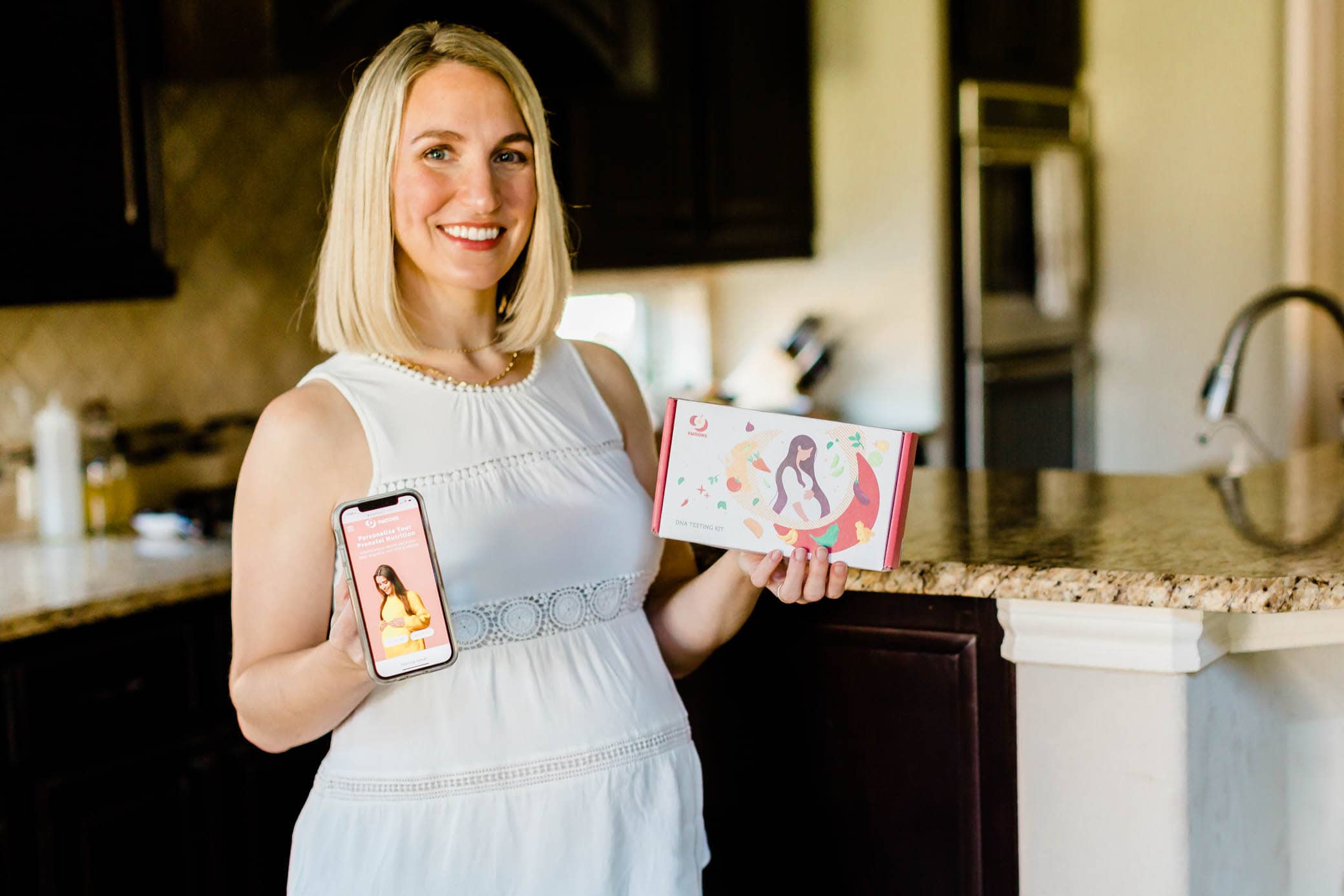 Pregnant mom standing in her kitchen holding up the 9moons app and DNA test kit.