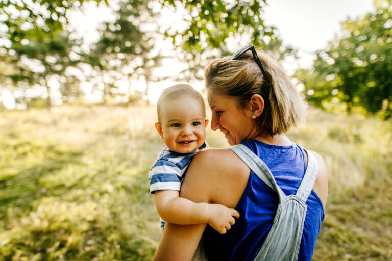 A woman carrying a smiling baby in an outdoor park captures a moment of pure joy. The baby, in a striped shirt, and the woman, in a blue tank top with sunglasses perched on her head, are surrounded by trees and grass on a sunny day. This is one of those moments you never apologize for cherishing.