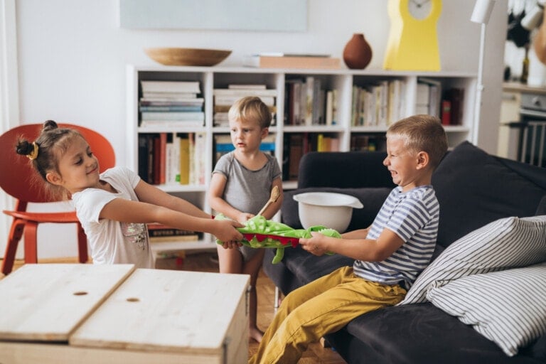 Three children are in a living room. Two are sitting on a couch tugging on a toy between them, while the third stands nearby watching with a toy dinosaur in hand. The room, furnished with bookshelves and a guitar, sets the scene for the kids learning to fight fair over their playthings.