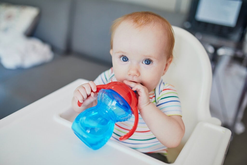 Little baby girl sitting in high chair at home and drinking water from a sippy cup.