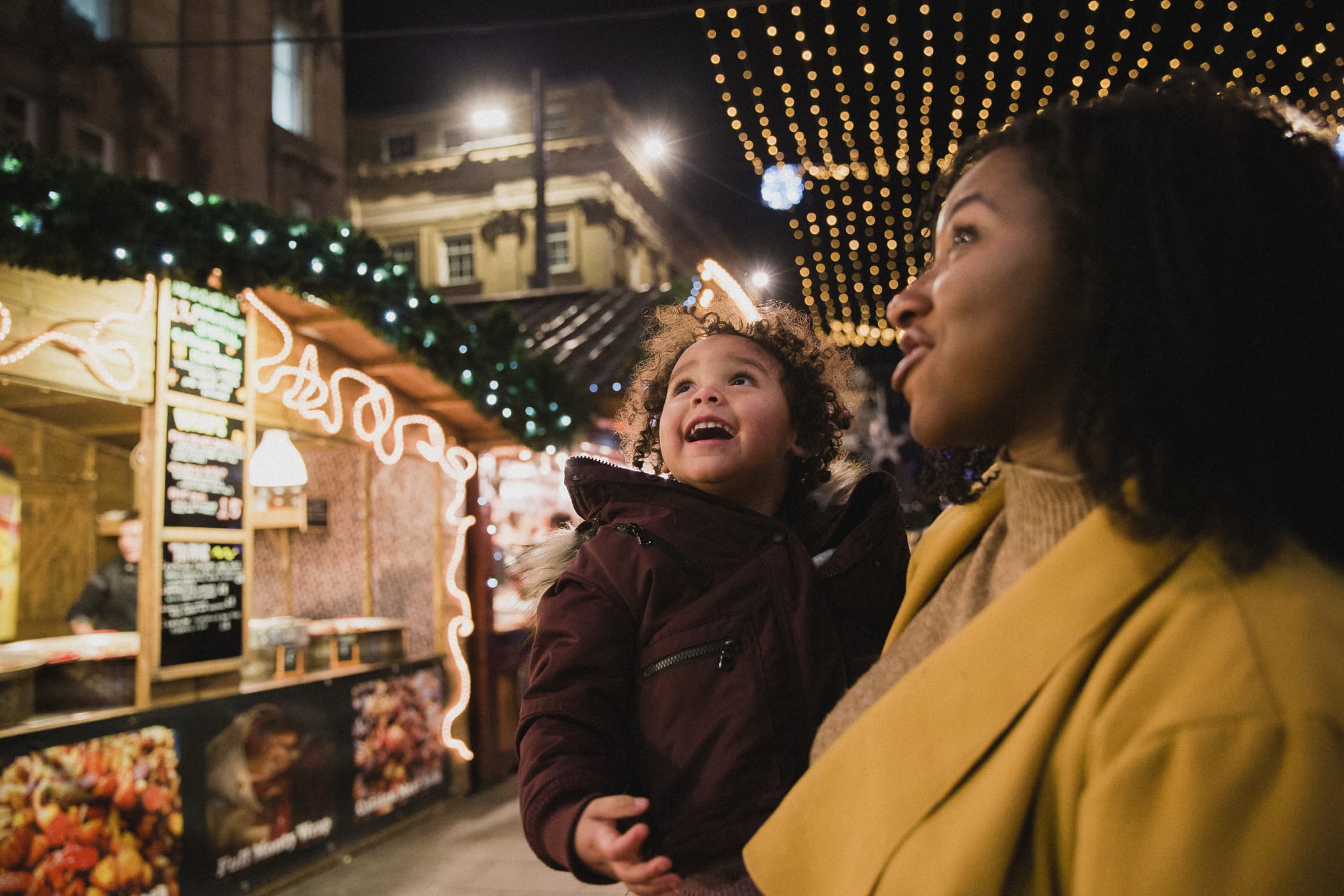 A close-up side-view shot of a woman holding her young daughter, they are both looking at bright​ Christmas lights at the Christmas markets on a cold night, the young girl is in awe.