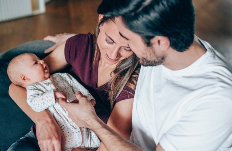 A couple is sitting closely on a couch, adoring their baby. The woman, wearing a maroon top, holds the baby who is looking up at the man. The man, in a white shirt, touches the baby's hand gently. They share a warm relationship in a softly lit room.