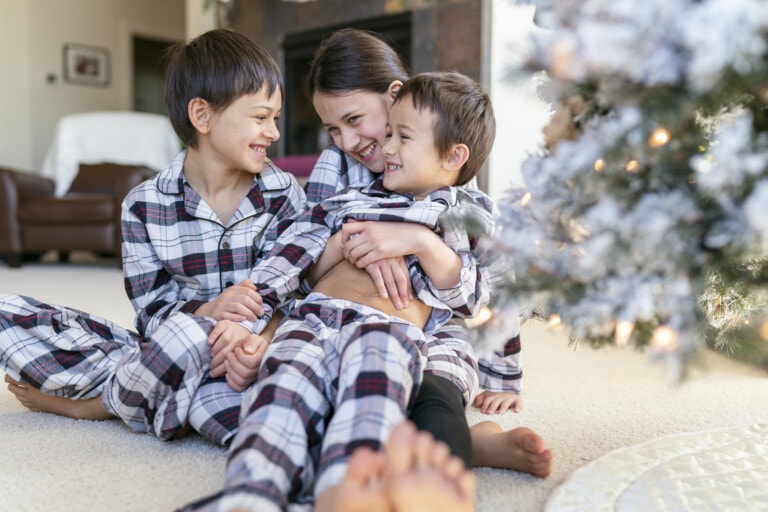Three mixed race Eurasian children wearing matching pajamas snuggle and laugh next to the Christmas tree set up in the living of their cozy home.