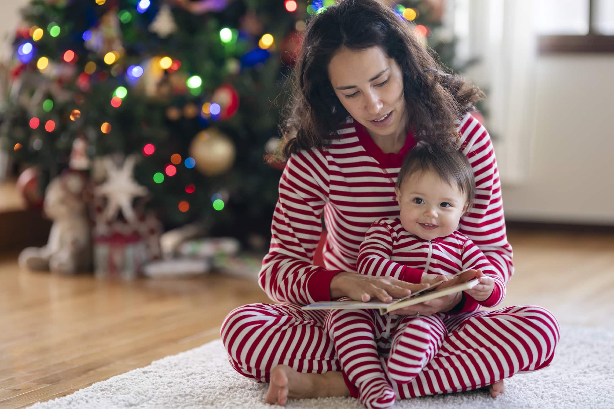 An adorable mixed race baby sits in her mother's lap and smiles as they read a book together. The family is celebrating the baby's first Christmas with a cozy day at home.