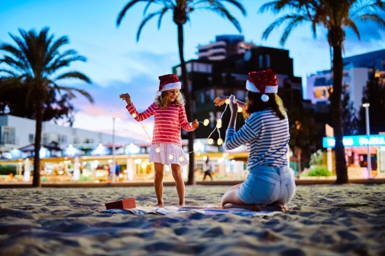 Two individuals, wearing Santa hats and striped shirts, are on a sandy beach, sharing holiday experiences. One is sitting and holding string lights, while the other stands, interacting with the lights. Palm trees and a modern cityscape are visible in the background at dusk.