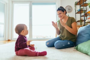 A woman is sitting on the floor clapping her hands and smiling at a baby in a maroon outfit, singing nursery rhymes. The baby is sitting across from her on a light-colored rug in a room with large windows and various plants.