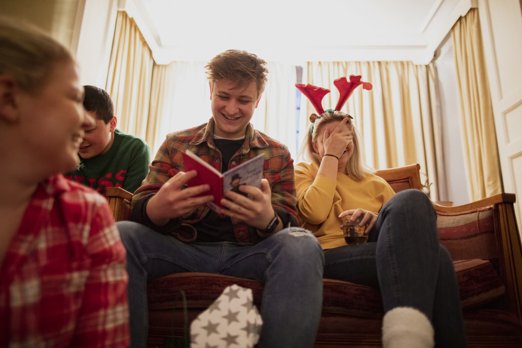 Young people are sitting together in the living room at a house party, opening Christmas presents.