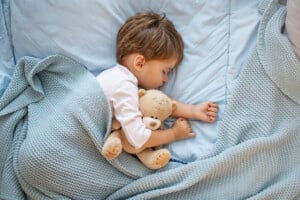 A young child with brown hair is sleeping on a bed with light blue sheets and a knit blanket. The child is hugging a brown teddy bear, one of their treasured loveys, lying on their side. The scene appears calm and quiet.