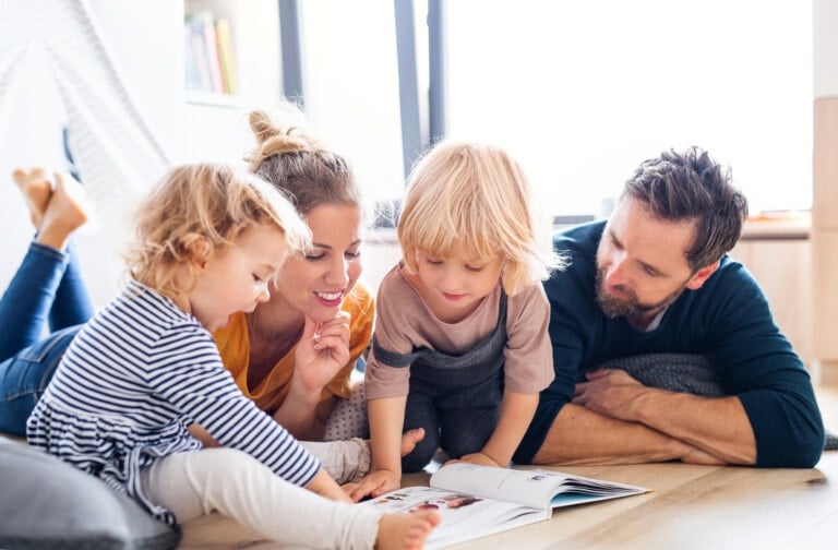 A blending family of four, including two adults and two young children, are lying on the floor in a bright, modern living room. They are engaged in reading a book together, with all members looking at the pages. The setting appears cozy and well-lit.