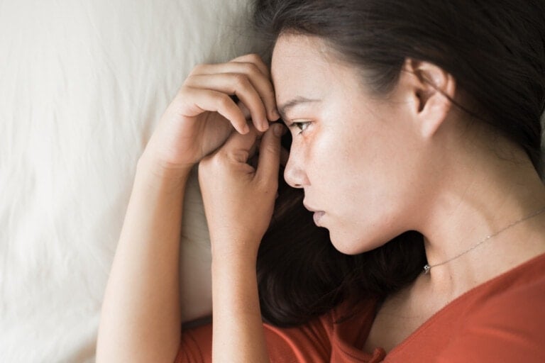 A woman wearing an orange top lies on her side on a white surface. Resting her head on her hand, she appears to be deep in thought, possibly reflecting on PMADs and their impact.