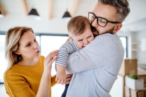 A woman in a yellow sweater gestures towards a man holding a crying toddler, perhaps indicating parental preference. The man, wearing glasses and a light blue shirt, is comforting the child. The background suggests a home setting with boxes visible, hinting at moving or unpacking.