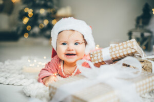 Little baby girl in Santa's hat is lying among the festive Christmas tree with presents.
