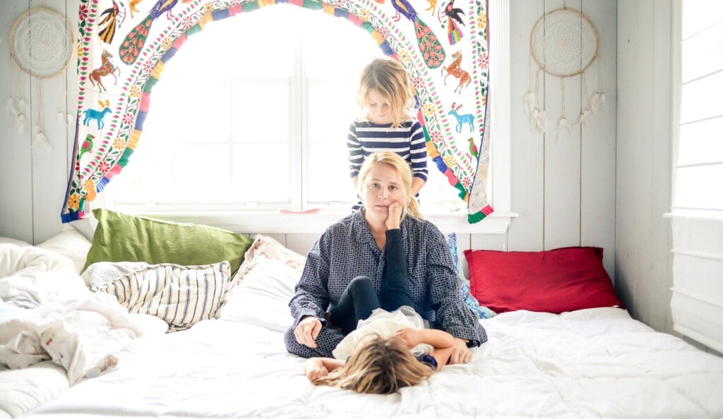 Mother in pajamas with coffee sits on the bed while children jump all around her