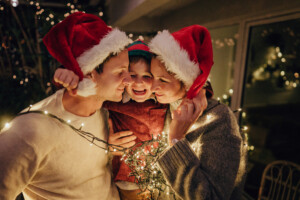 Photo of a family with one child wrapped in Christmas lights, celebrating holidays on the balcony of their apartment.