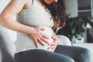 A person wearing a white sleeveless top and grey pants is holding their pregnant belly while sitting on a chair, contemplating the upcoming journey of uterine involution. The background includes a blurred plant and other household items out of focus.