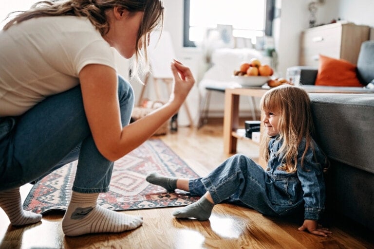 A woman squats on the floor, talking to a little girl who is sitting on the ground indoors. The girl is smiling and wearing a denim outfit. The room has wooden floors, a patterned rug, and a bowl of fruit on a table in the background, fostering an atmosphere free from any shaming.
