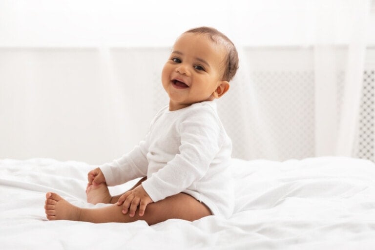 Portrait Of Adorable African American Baby Toddler Sitting On Bed And Smiling Looking At Camera Posing In Bedroom At Home.