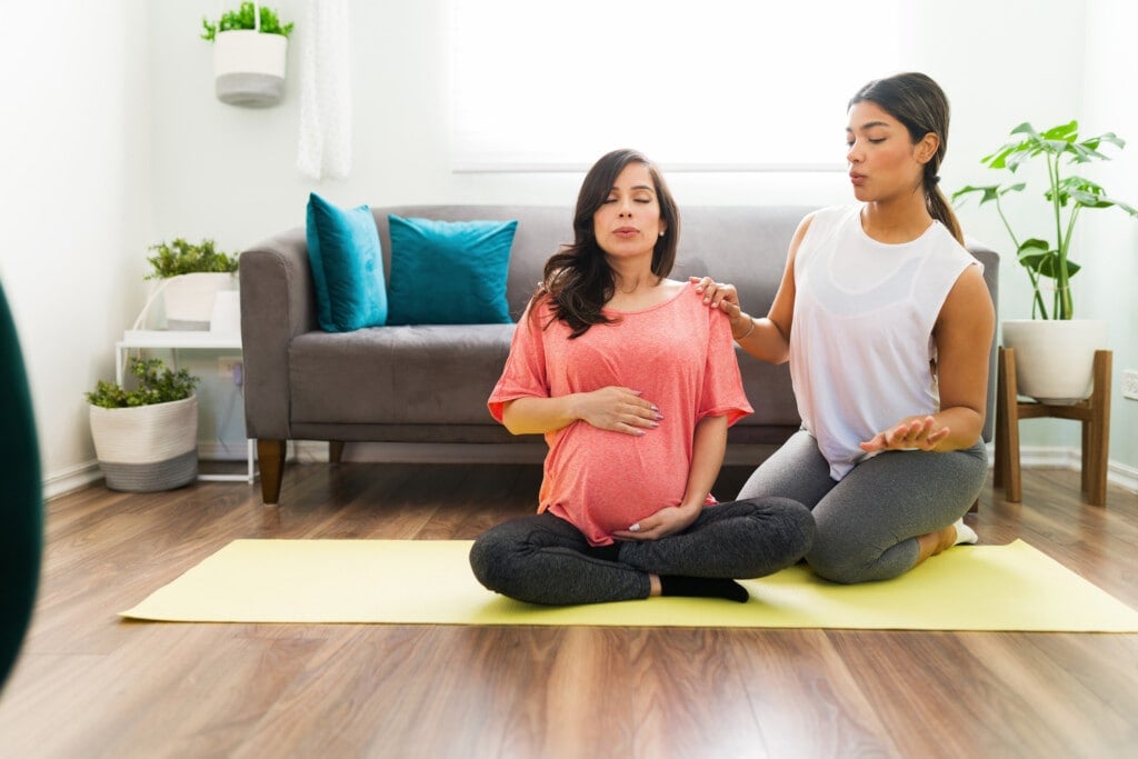 Sporty doula doing breathing exercises with a caucasian pregnant woman while sitting on a yoga mat in the living room