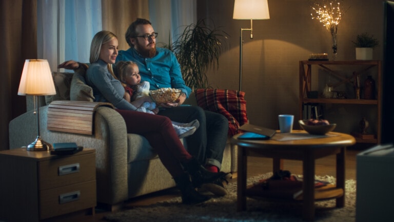 Long Shot of a Father, Mother and Little Girl Watching TV. They Sit on a Sofa in Their Cozy Living Room and Eat Popcorn. It's Evening.