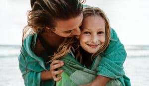 An adult hugs and kisses a smiling child on the cheek at the beach. Both are wrapped in green towels. The ocean is visible in the background.