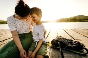 A woman and her son sit closely on a wooden dock by the water at sunset. The mother, wearing a white polka-dot shirt and green pants, embraces her smiling son, who is wearing a white shirt and green shorts. A black backpack rests nearby.