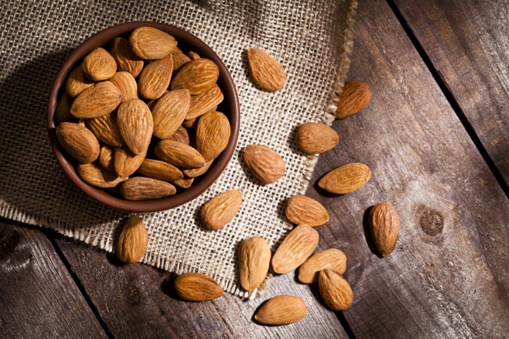 Top view of a brown bowl filled with organic almonds shot on rustic wood table. Some almonds are out of the bowl on a burlap.