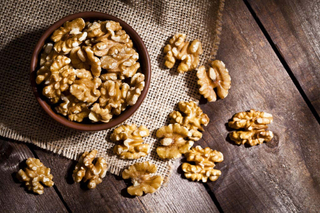 Top view of a brown bowl filled with organic walnuts shot on rustic wood table. Some almonds are out of the bowl on a burlap.