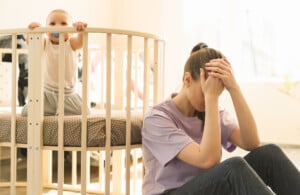 A woman sits on the floor with her head in her hand, looking stressed, while a baby stands in a crib and looks toward her.