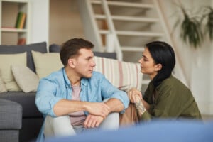 A man and a woman sit on the floor in a living room, facing each other and engaged in conversations. The man wears a light blue shirt and jeans, while the woman wears a green top. They appear to be in a relaxed home setting with a couch and a staircase in the background.