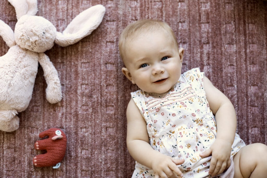 Directly above view of cute baby girl lying on carpet. Portrait of female toddler is by stuffed toy animals. She is wearing floral dress in living room.