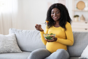 Healthy diet during pregnancy. Smiling pregnant black woman enjoying fresh vegetable salad at home. Expecting african american lady sitting on sofa at living room and eating healthy food.