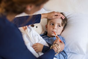 Sick boy with thermometer laying in bed and mother hand taking temperature. Mother checking temperature of her sick son who has thermometer in his mouth. Sick child with fever and illness while resting in bed.
