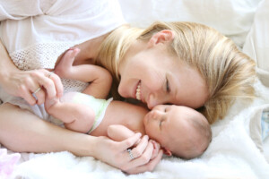 A happy young mother is laying in her white bed, lovingly hugging and snuggling her newborn baby daughter.