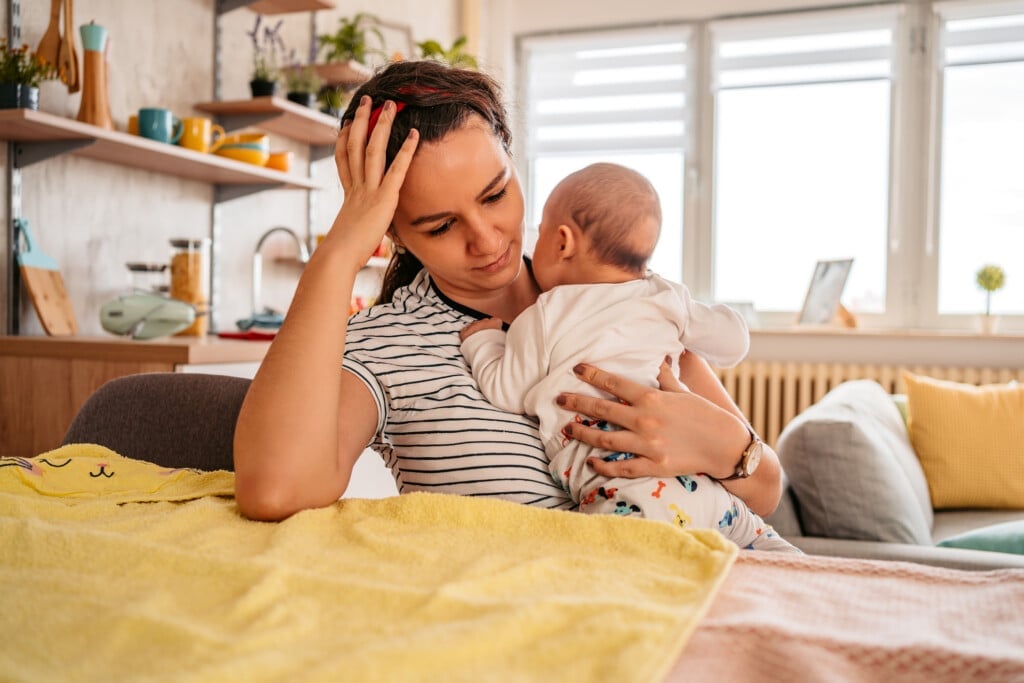 Young adult mother sitting in chair at home, holding her three month old baby son, feeling tired, sleepy or having a headache.