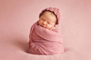 picture of a newborn baby girl swaddled in a pink muslin swaddle with a pink background. The photo is taken in a studio.