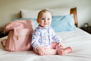 Baby boy sitting on a big bed with a pink diaper bag next to him.